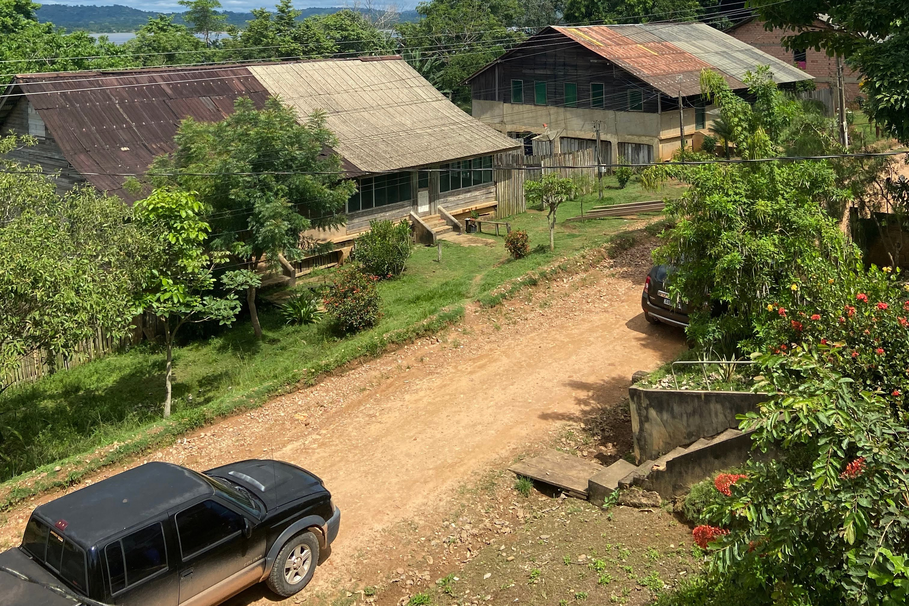 Rural farm compound with multiple buildings, dirt driveway, parked vehicle, and surrounding green vegetation on hillside landscape.