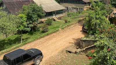 Rural farm compound with multiple buildings, dirt driveway, parked vehicle, and surrounding green vegetation on hillside landscape.
