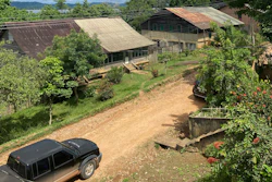 Rural farm compound with multiple buildings, dirt driveway, parked vehicle, and surrounding green vegetation on hillside landscape.