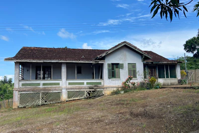 Weathered white colonial-style house with dark roof and lattice foundation on rural hillside