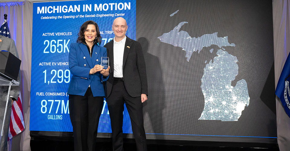 Two professionals stand at a podium during Michigan In Motion event celebrating Social Engineering Center opening, with vehicle statistics displayed on blue banner.