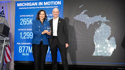 Two professionals stand at a podium during Michigan In Motion event celebrating Social Engineering Center opening, with vehicle statistics displayed on blue banner.