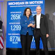 Two professionals stand at a podium during Michigan In Motion event celebrating Social Engineering Center opening, with vehicle statistics displayed on blue banner.