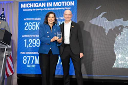 Two professionals stand at a podium during Michigan In Motion event celebrating Social Engineering Center opening, with vehicle statistics displayed on blue banner.