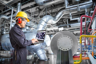 Industrial technician monitoring HVAC system with laptop in factory facility with large turbine equipment