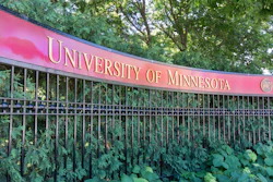 University of Minnesota pink banner sign mounted on decorative fence with green foliage backdrop