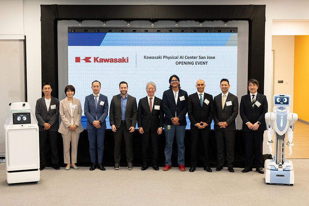 Ten business professionals stand in formal attire at Kawasaki Physical AI Center San Jose opening event, flanked by robotic equipment.