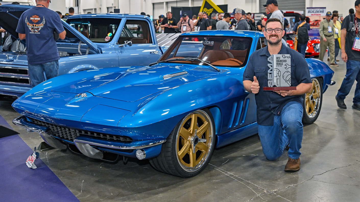 Man posing with blue 1965 Chevrolet Corvette Stingray with gold wheels at indoor car show