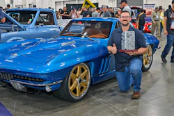 Man posing with blue 1965 Chevrolet Corvette Stingray with gold wheels at indoor car show