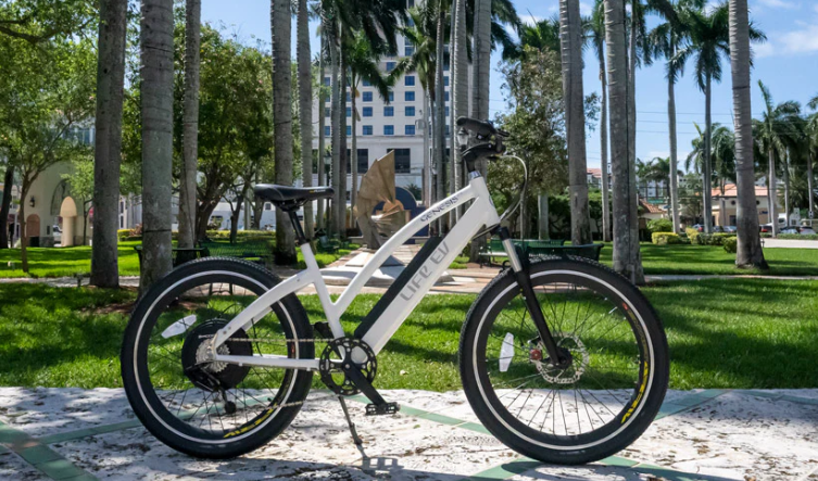 White and black hybrid bicycle displayed on a palm tree-lined path with modern buildings in the background