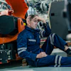 Tired industrial worker in blue safety suit sitting on floor next to orange machinery in factory setting