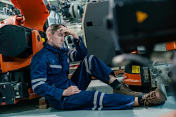 Tired industrial worker in blue safety suit sitting on floor next to orange machinery in factory setting