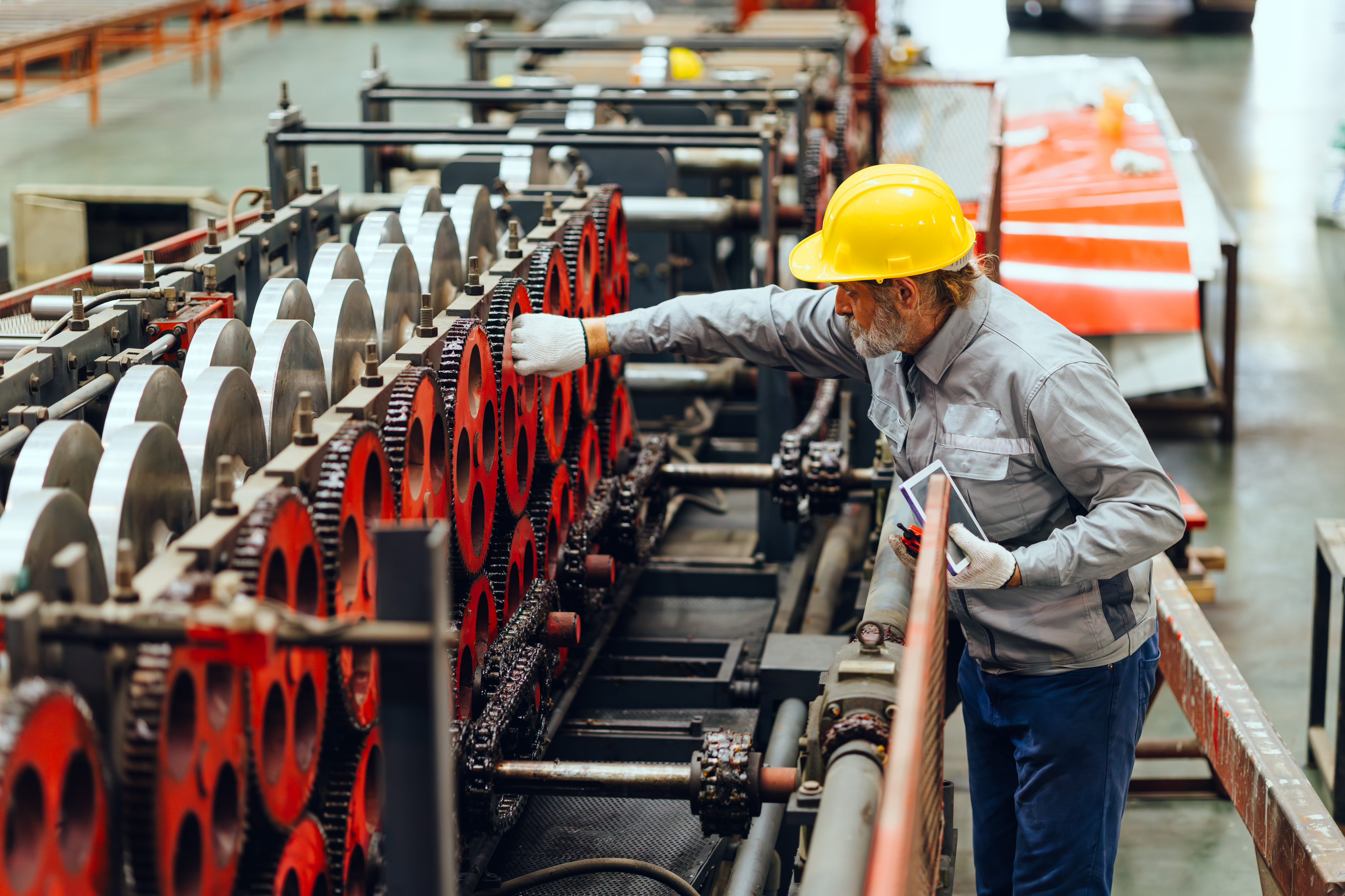 Factory worker in yellow hard hat inspecting red industrial machinery gears in manufacturing facility
