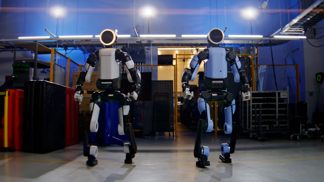 Two white and blue humanoid robots standing in an industrial warehouse facility with overhead lighting and equipment