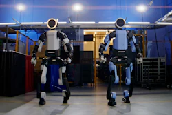 Two white and blue humanoid robots standing in an industrial warehouse facility with overhead lighting and equipment
