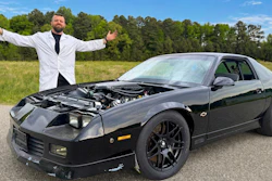 Man in white lab coat posing beside black Pontiac Firebird with open hood displaying engine