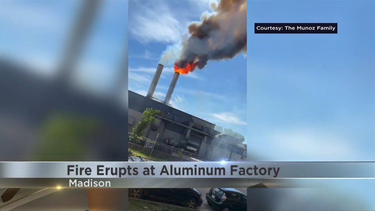 Fire erupts from industrial aluminum factory with twin smokestacks in Madison, thick black smoke visible against blue sky