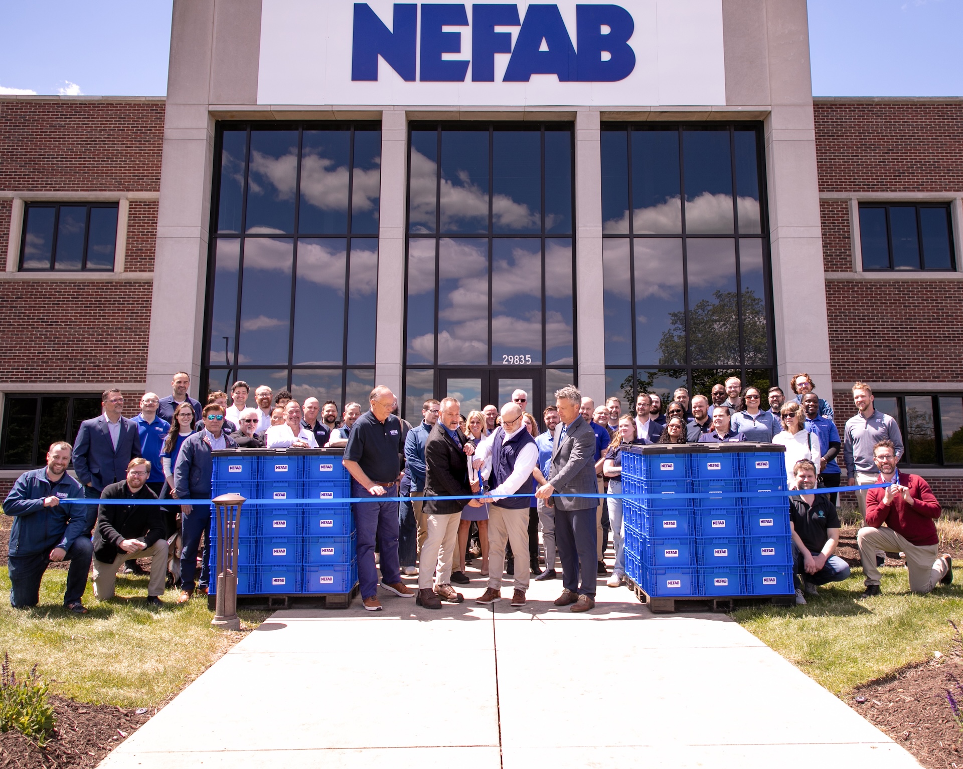 Large group of NEFAB employees gathered for ribbon-cutting ceremony in front of company headquarters with blue crates