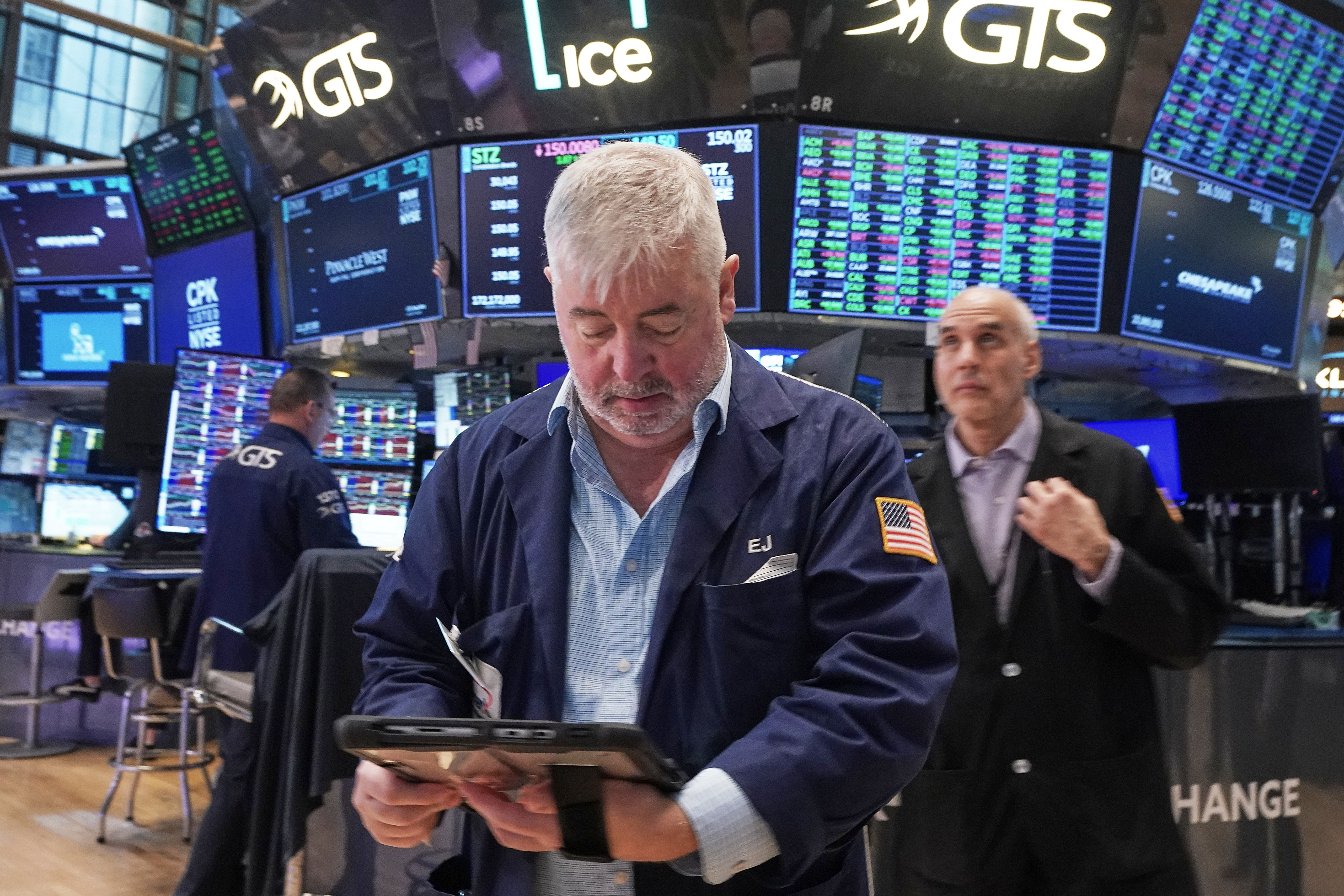 Trader in navy jacket examines tablet at stock exchange trading floor with multiple monitors displaying market data