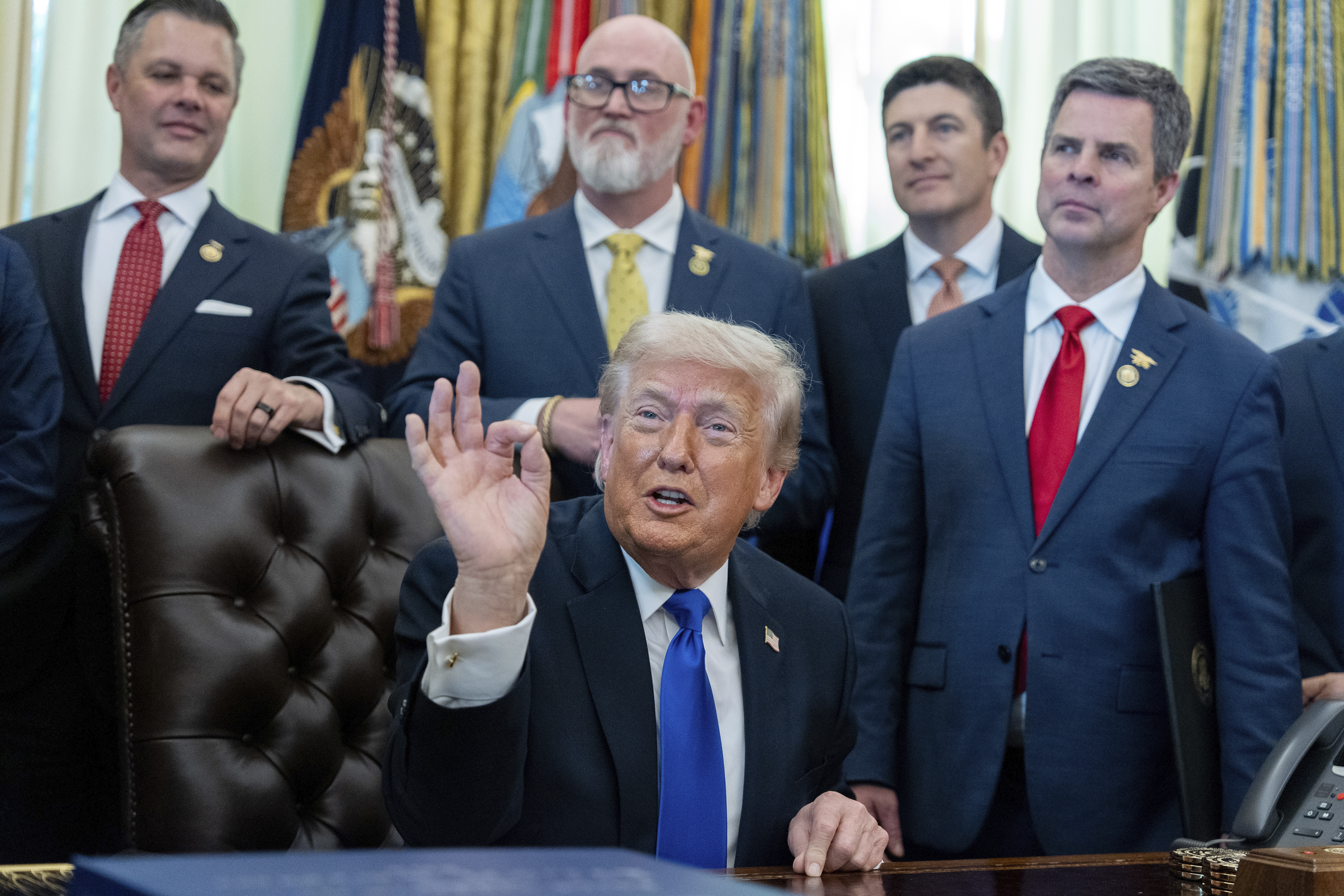 Five men in business suits stand in formal office setting with American flags and ornate drapes in background