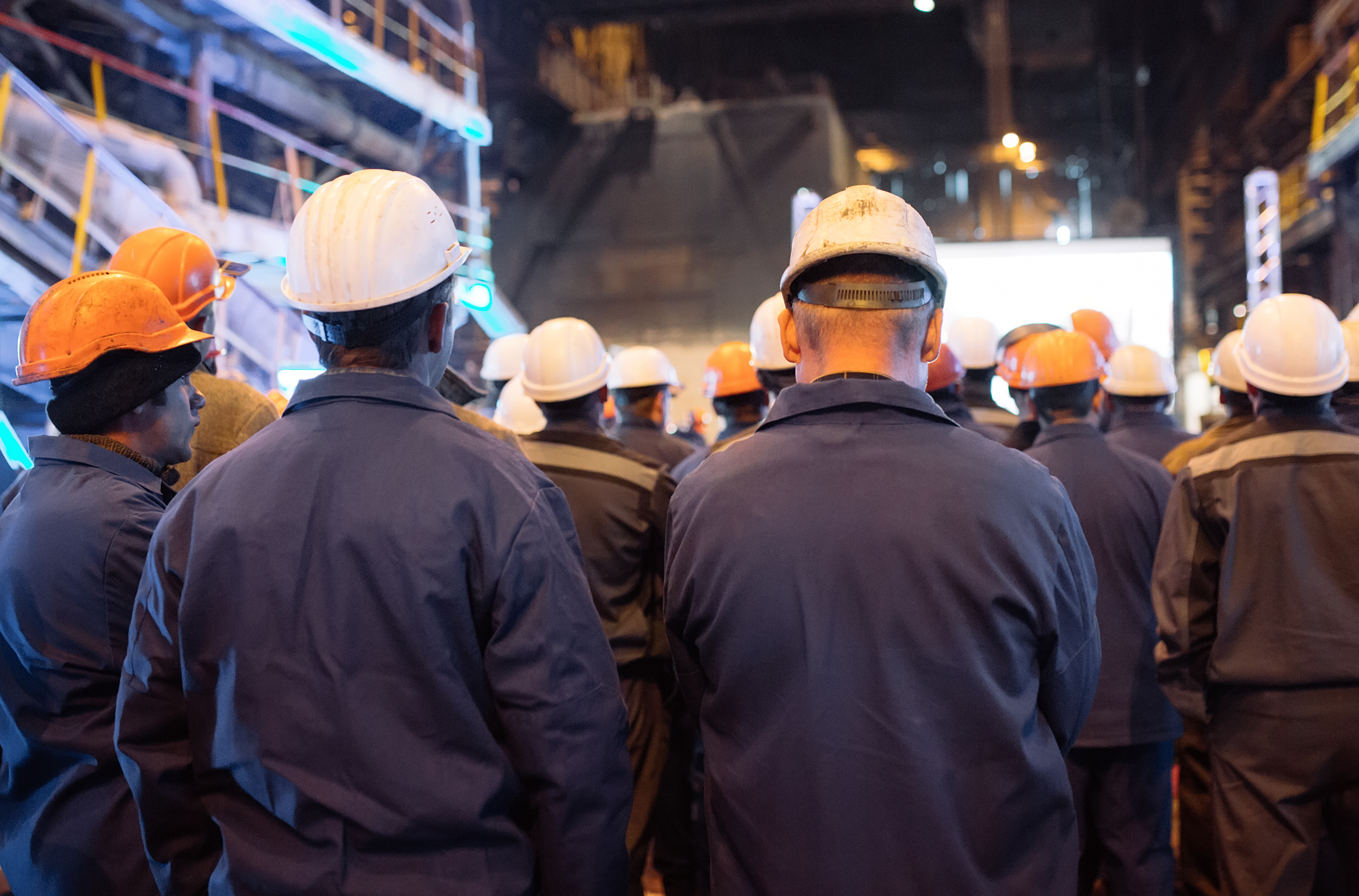 Construction workers wearing safety helmets and dark jackets gathered at a nighttime job site with illuminated buildings in the background.