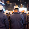 Construction workers wearing safety helmets and dark jackets gathered at a nighttime job site with illuminated buildings in the background.