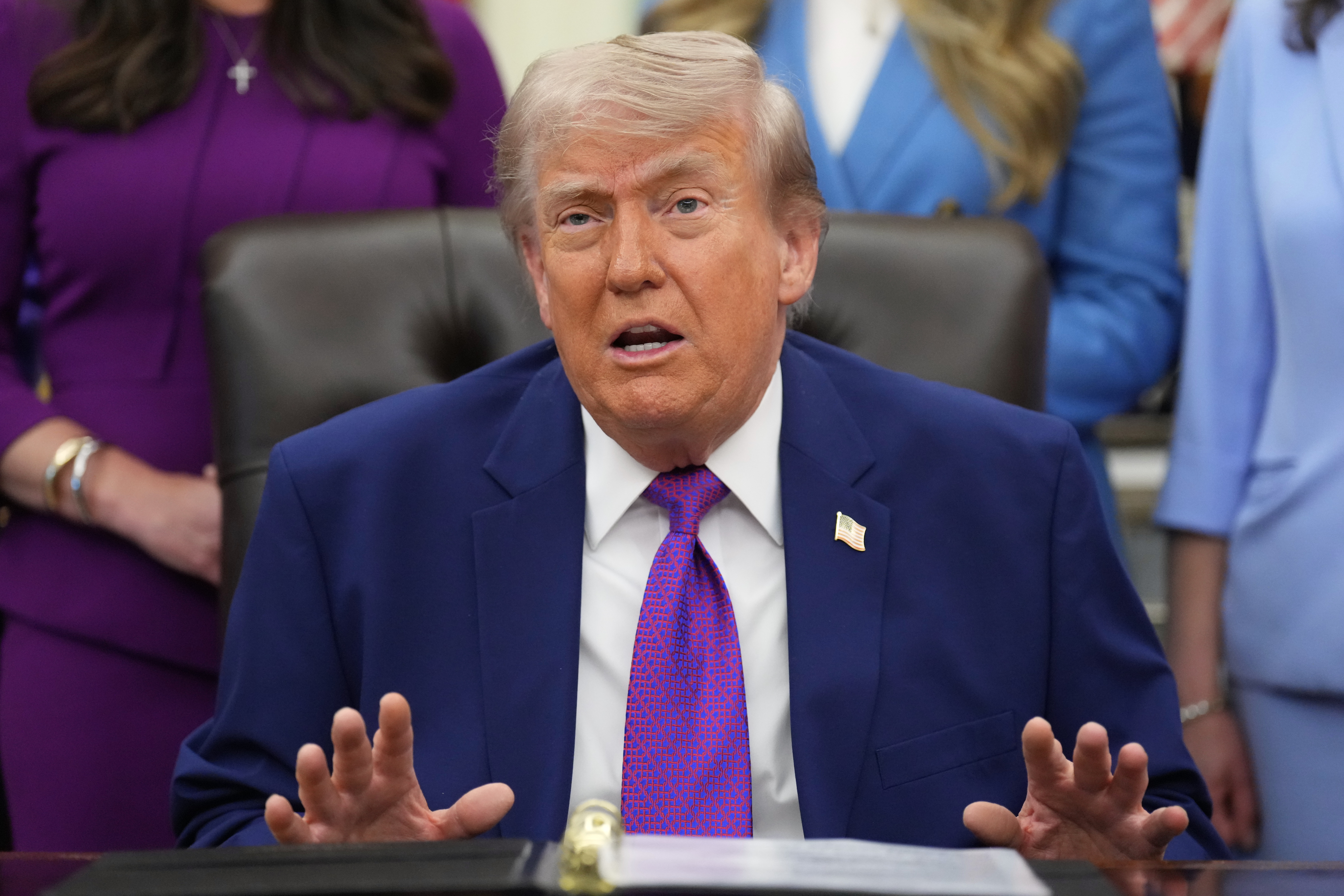 Man in dark blue suit and purple tie speaking at formal desk with attendees in background