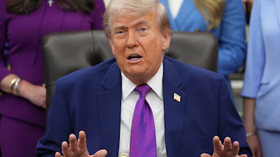 Man in dark blue suit and purple tie speaking at formal desk with attendees in background