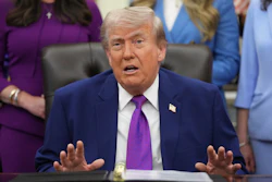 Man in dark blue suit and purple tie speaking at formal desk with attendees in background