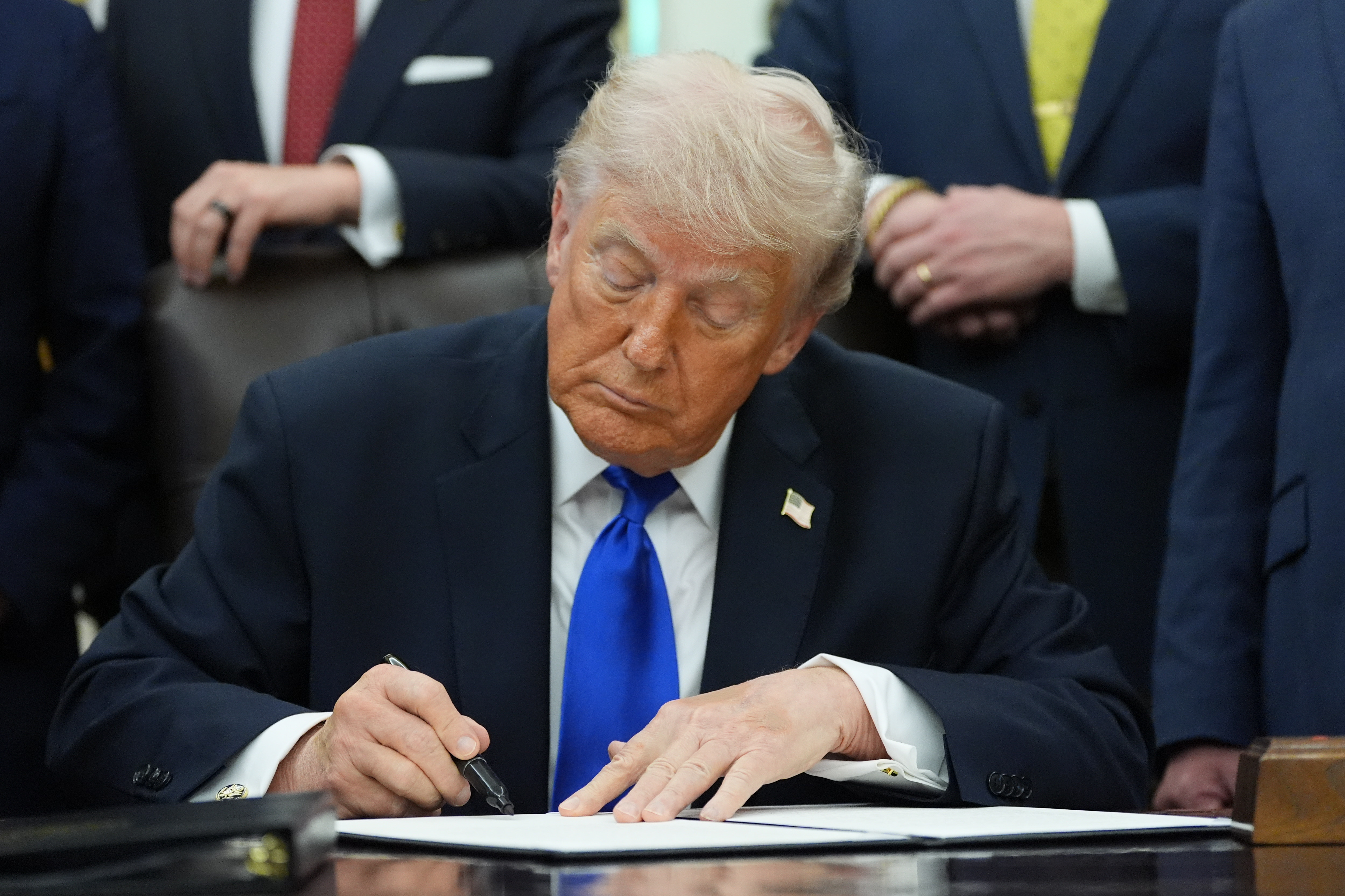 Man in dark suit and blue tie signing document at desk surrounded by officials