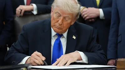 Man in dark suit and blue tie signing document at desk surrounded by officials