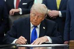 Man in dark suit and blue tie signing document at desk surrounded by officials