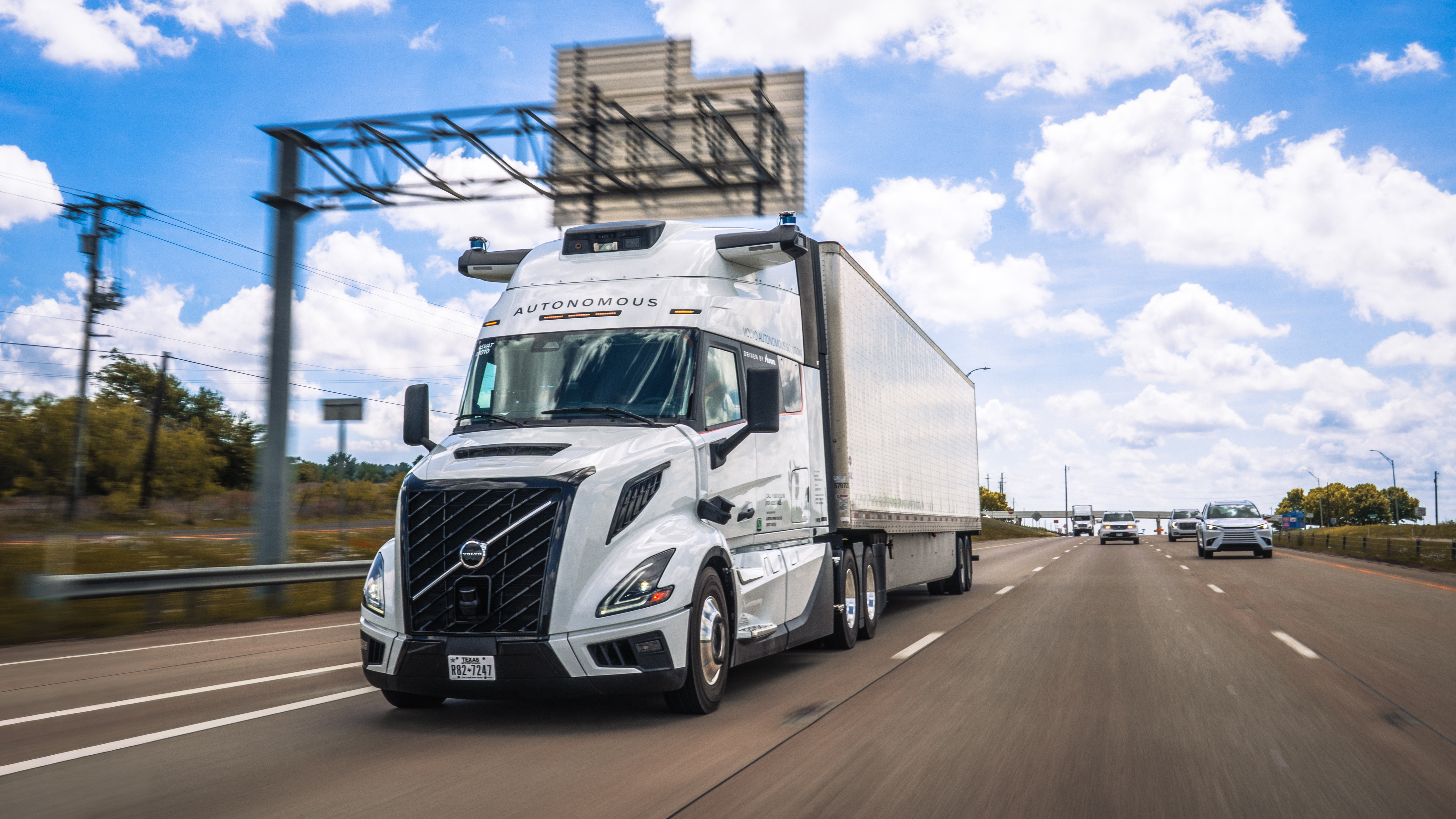 White Volvo autonomous semi-truck driving on highway with clear blue sky and traffic
