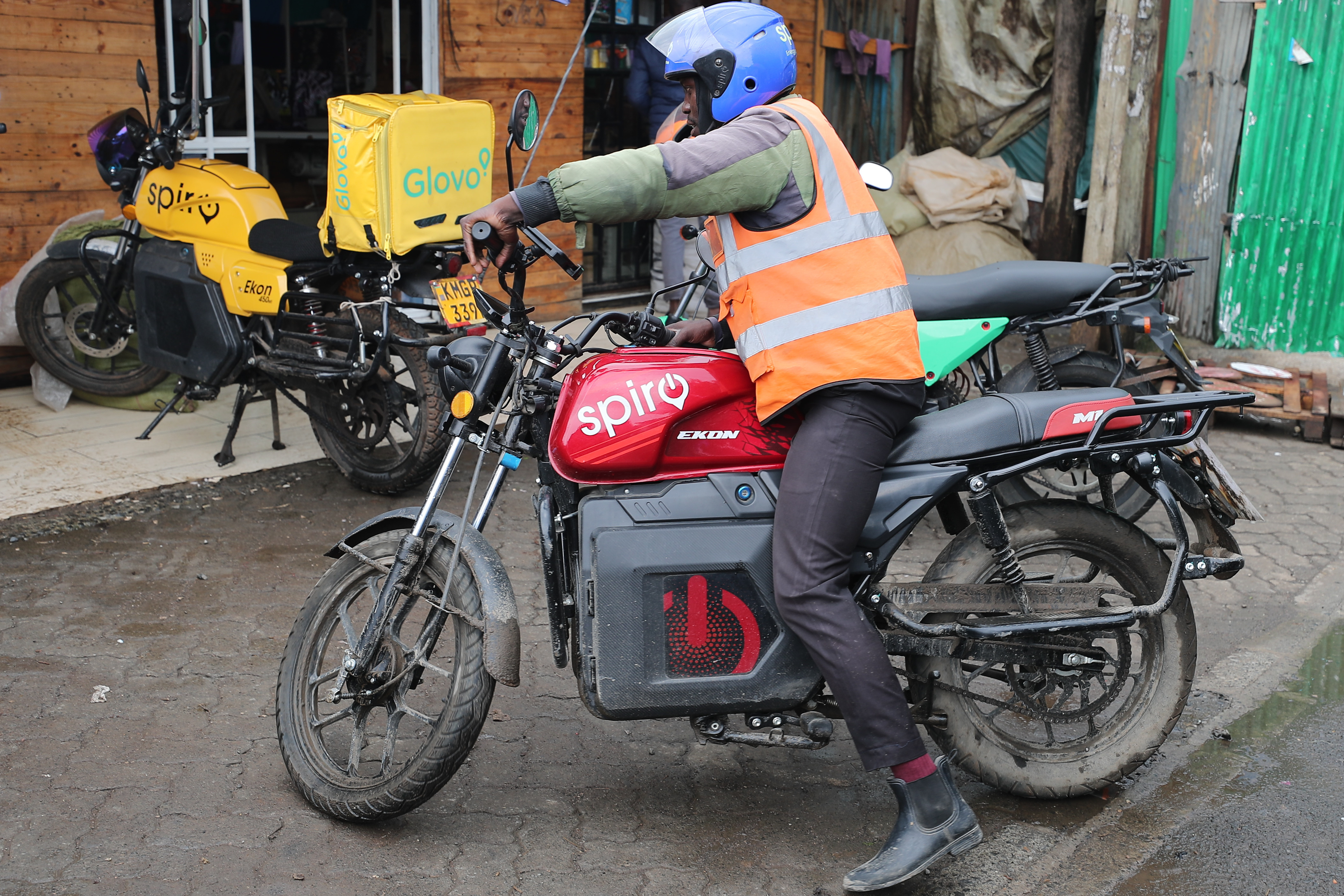 Delivery worker in orange safety vest and blue helmet sitting on red Spiro motorcycle with delivery box in urban setting