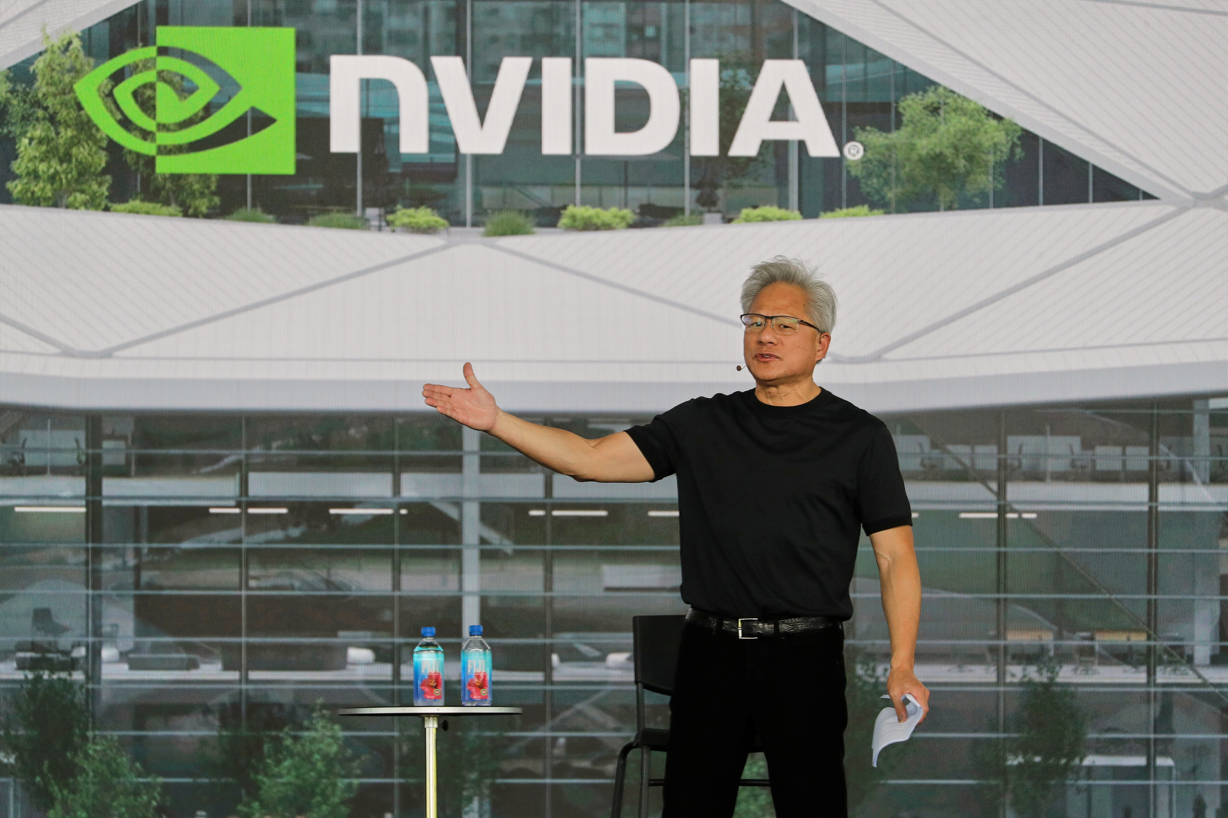 Man in black shirt speaking at NVIDIA corporate headquarters with green and white logo visible on building backdrop
