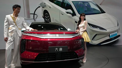 Two people in formal attire stand beside a red electric sedan at an auto showroom with white vehicles in the background.