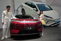 Two people in formal attire stand beside a red electric sedan at an auto showroom with white vehicles in the background.
