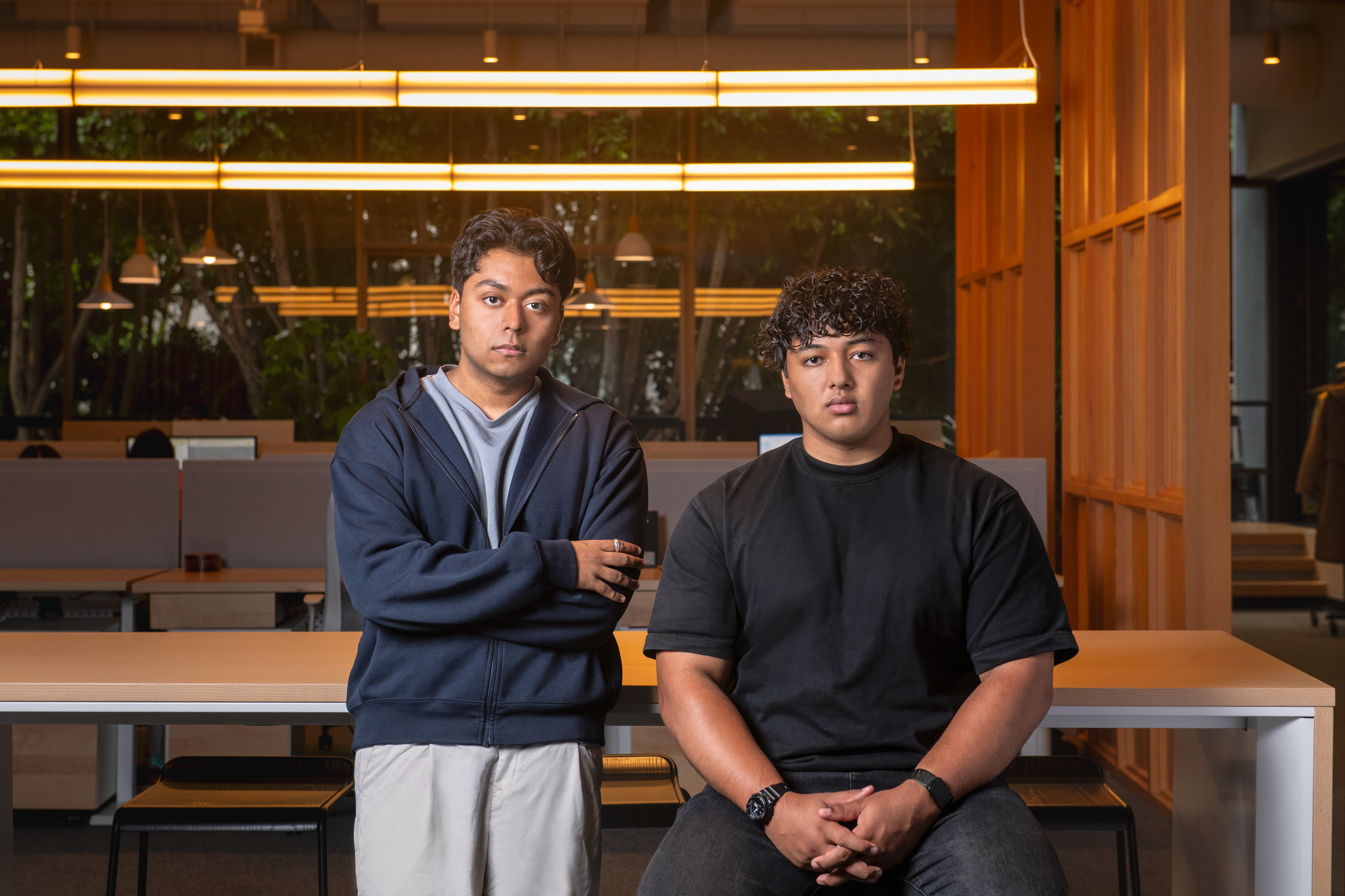 Two young men sitting on a bench in a modern office lobby with warm lighting and wooden architectural elements.