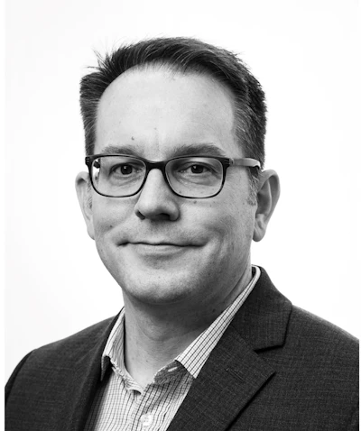 Professional headshot of a man wearing glasses and dark blazer with checkered shirt