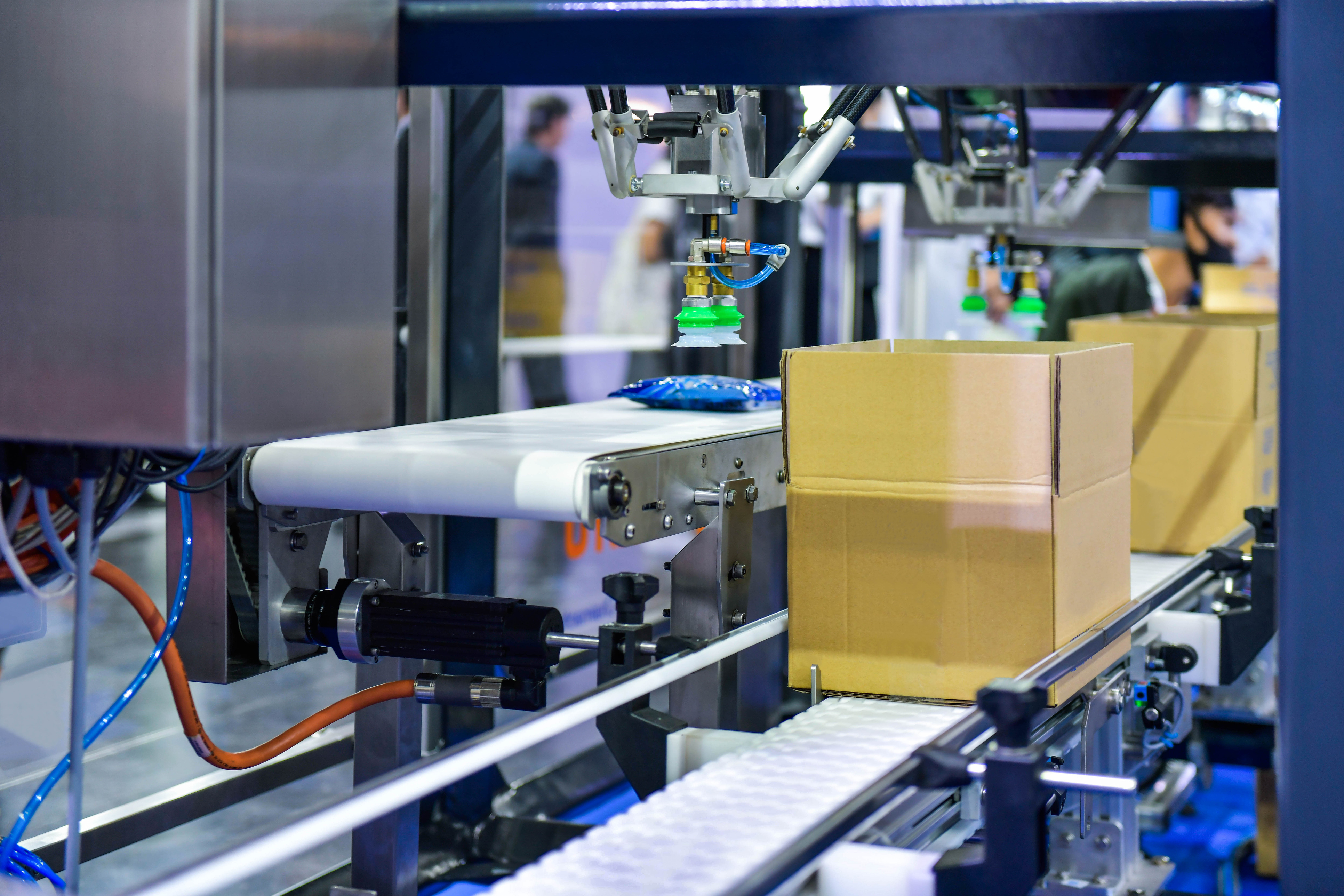 Industrial 3D printer or automated manufacturing system with yellow cardboard boxes on conveyor belt and printing head in operation