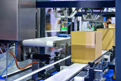 Industrial 3D printer or automated manufacturing system with yellow cardboard boxes on conveyor belt and printing head in operation