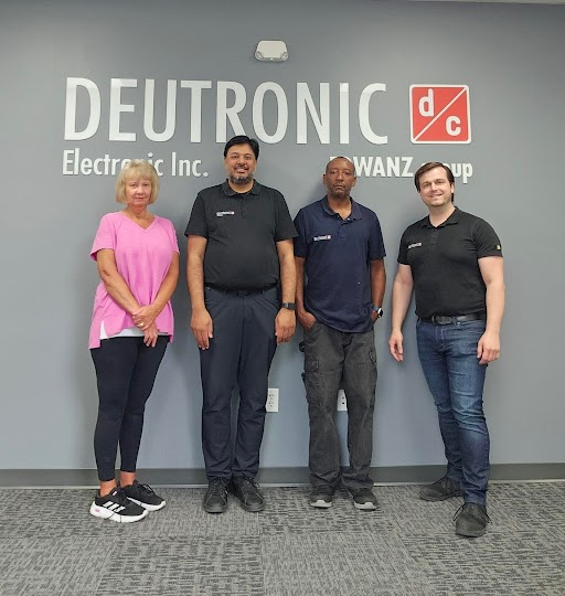 Four team members posing in front of Deutronic Electronics Inc. corporate office wall signage