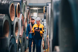 Two industrial workers in safety gear inspecting stacked steel coils in a warehouse facility