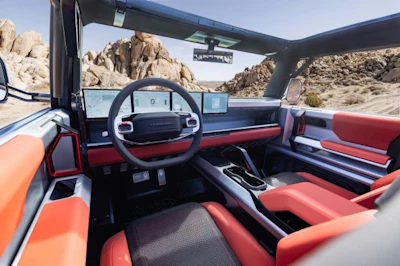 Interior view of a convertible vehicle with red and black upholstery, steering wheel, and dashboard overlooking a desert landscape with rocky terrain.