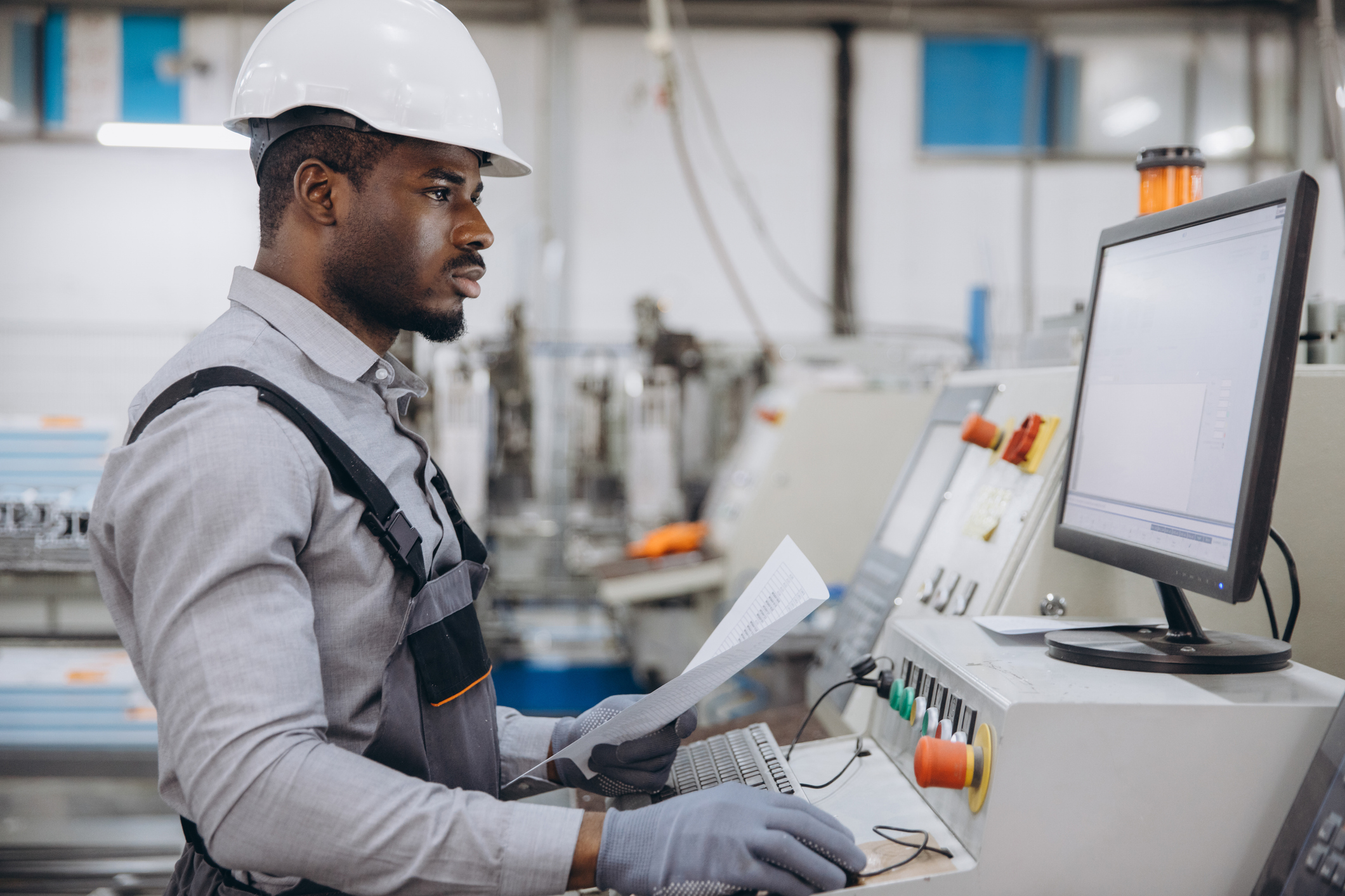 Industrial worker in hard hat reviewing documents at factory control panel with monitor