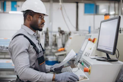 Industrial worker in hard hat reviewing documents at factory control panel with monitor