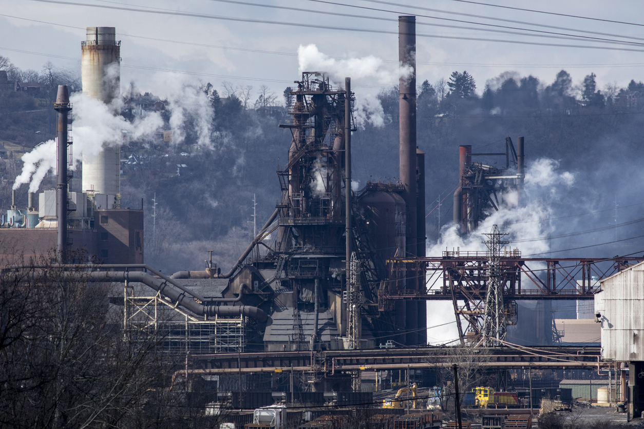 Industrial steel mill facility with multiple smokestacks and processing towers emitting steam against a forested hillside backdrop.