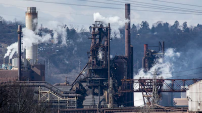 Industrial steel mill facility with multiple smokestacks and processing towers emitting steam against a forested hillside backdrop.