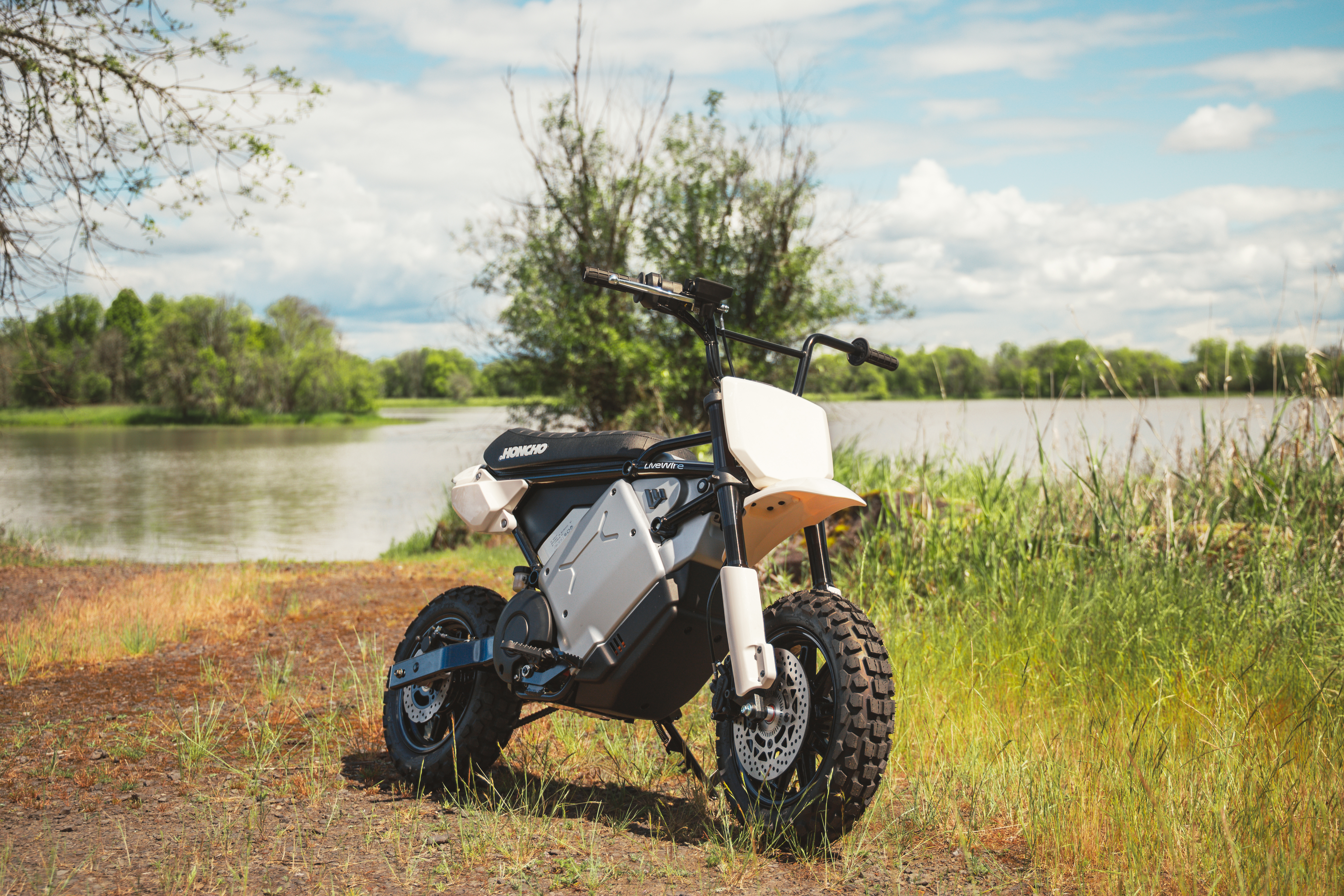 Orange and white electric scooter parked on grass near a river with trees and blue sky