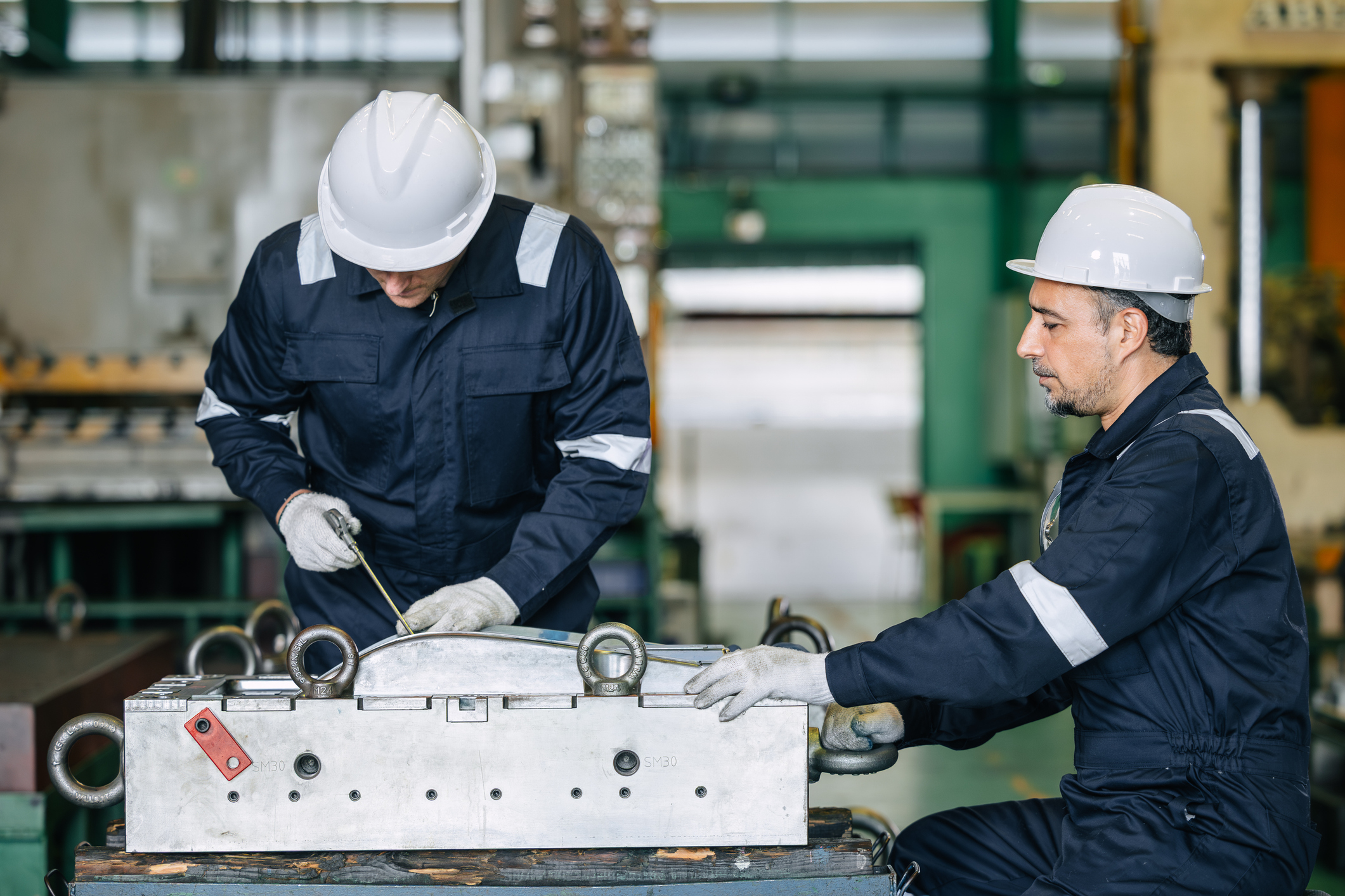 Two industrial workers in hard hats and safety gear inspect equipment in a manufacturing facility.